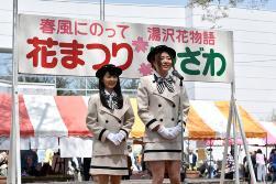 「花まつり ゆざわ」と書かれてある横断幕の前で、黒い帽子に胸にはチェックのリボン、クリーム色のお揃いのジャケットにミニスカートの服を着た若い女性2名がスタンドマイクの前に立って話をしている写真