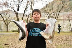 黒いティーシャツを着た男の子がビニール袋に大きなニジマスを1匹ずつ入れて両手に持って見せている写真