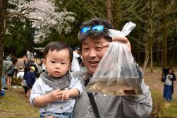 小さな男の子を抱っこした男性が、水の入ったビニール袋の中の生きたニジマスを持って見せている写真