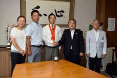 オレンジ色のリボンのメダルを首から下げた今村 豪選手と町長が握手をしており、関係者の方々と一緒に写っている写真