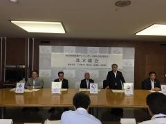 魚沼圏域版新ナンバープレート導入実行委員会設立総会で、並んで着席する男性と、その中で立って話をしている男性の写真