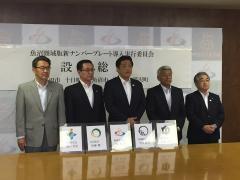魚沼圏域版新ナンバープレート導入実行委員会設立総会で、並んでいる5名の男性と、その前に並べられたアクリルフレームに入った5枚の書類の写真