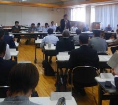 会議で、着席して話を聞く参加者と、前方で立って話をする男性の写真