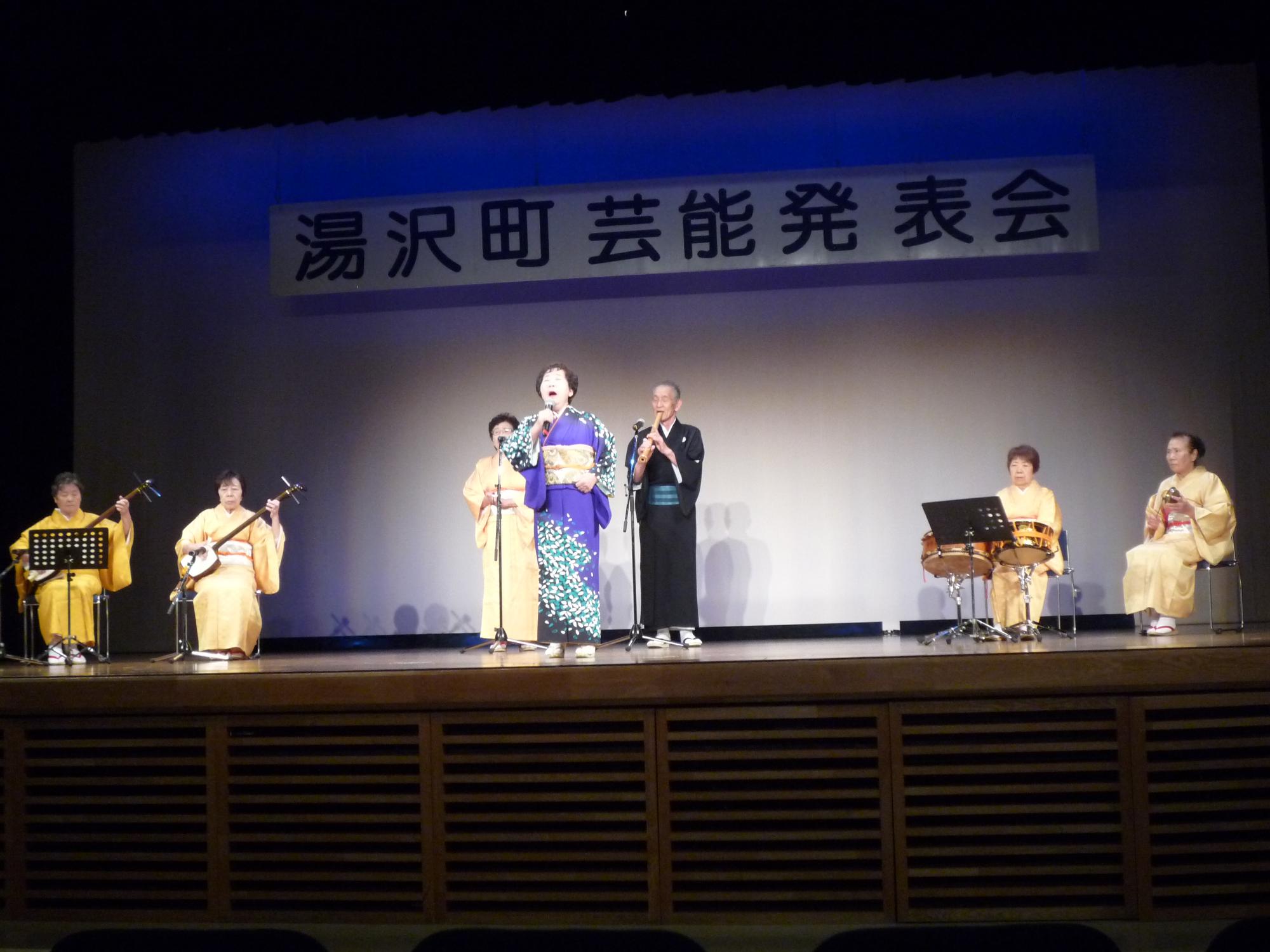 舞台で民謡を歌唱、演奏するのすみれ会のメンバーの写真