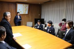 大きなテーブルの席に長谷川選手、金井選手、太田選手が座っており、町長が3選手の前に立って話をしている写真