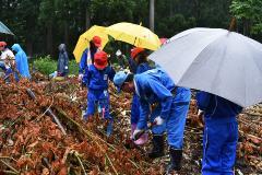 スコップを持った男性が土を掘っており、雨の中、傘をさしながら、周りの児童がその様子を見ている写真