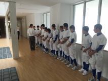 白いユニフォームを着た湯沢学園野球部員が廊下に一列に整列しており、今村 豪選手が部員の皆さんと話をしている写真