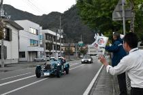 水色の車体のクラシックカーが沿道を走っており、役場前で職員の方々が旗を持って、通り過ぎるクラシックカーに手を振っている写真