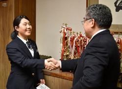 優勝トロフィーが沢山並んでいる台の前で、金井選手と田村町長が握手を交わしている写真