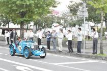 水色の車体のクラシックカーが沿道を走っており、役場前に職員の方々が立って通り過ぎるクラシックカーを見て手を振っている写真