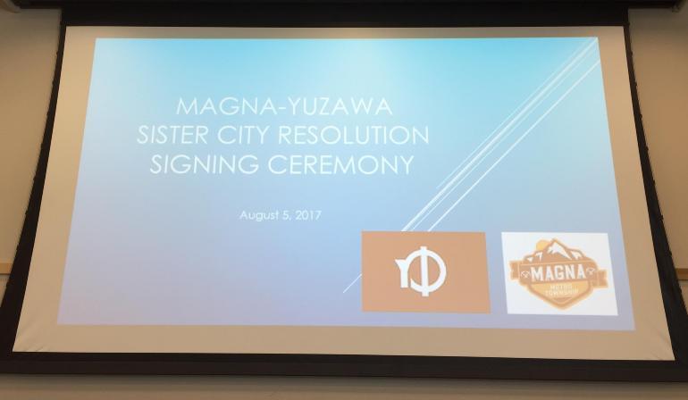 プロジェクタースクリーンに映る英語で書かれたマグナメトロタウンシップとの姉妹都市協定調印式の写真